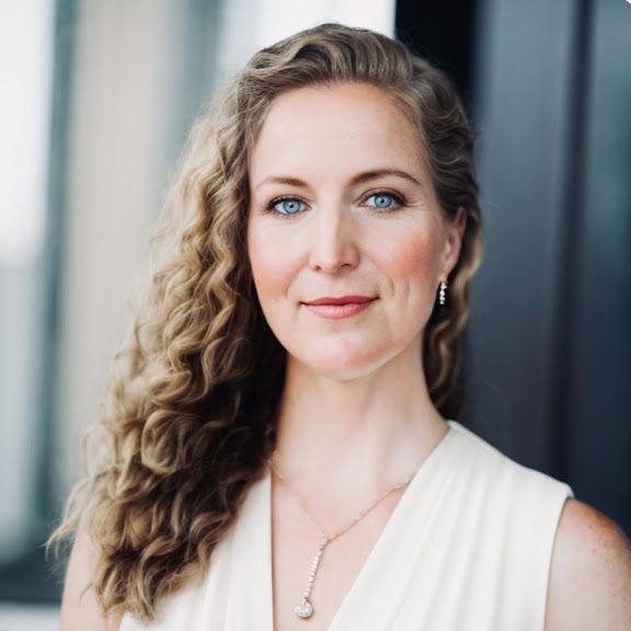 Person with long curly hair wearing a cream sleeveless top and a necklace with a pendant.
